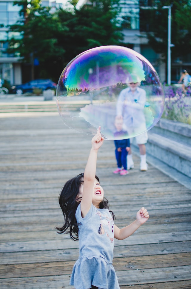 Ein kleines Mädchen piekst neugierig und lachend in eine große Seifenblase